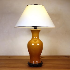 Può includere: Lampada da tavolo decorativa con paralume bianco e base in ceramica color oro. La base ha una forma a vaso e poggia su una base circolare nera. La lampada è su una superficie di legno.