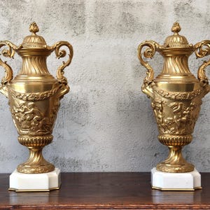 May include: A pair of ornate gold-tone vases with marble bases. Each vase features intricate details, including a sculpted design of figures and foliage.