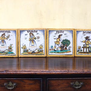 May include: Four square ceramic tiles illustrating the seasons: Primavera, Verano, Otoño, and Invierno. Each tile shows a person in period attire, with floral and natural motifs. The tiles are set in a wooden frame and are displayed on a dark wooden surface.