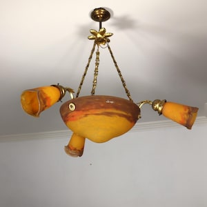 May include: An antique ceiling light fixture with a unique design. The fixture features a central, oval-shaped body in shades of yellow and orange, with three trumpet-shaped lamps. The fixture is suspended by brass chains.