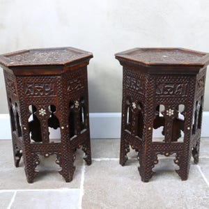 Può includere: Due tavolini in legno marrone scuro, intagliati in modo intricato. Ogni tavolo ha un piano esagonale e lati traforati ornati con motivi floreali e geometrici. I tavoli sono autoportanti e hanno gambe decorative.