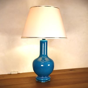 Può includere: Una lampada da tavolo in ceramica turchese con un paralume bianco. Il paralume ha un bordo dorato intorno alla parte superiore e inferiore.