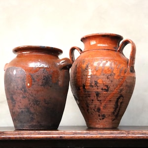 Può includere: Due antichi vasi in terracotta dall'aspetto rustico e invecchiato. I vasi sono marroni con segni più scuri e macchiati e una finitura lucida. Uno ha una forma arrotondata, l'altro è più alto con manici.