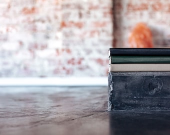 May include: A stack of three notebooks on a concrete block. The notebooks are black, green, and beige. The background is a blurred brick wall.