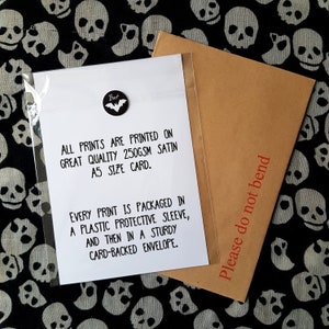 May include: A white card with black text that reads "All prints are printed on great quality 250gsm satin A5 size card. Every print is packaged in a plastic protective sleeve, and then in a sturdy card-backed envelope." The card is sitting on a black and white fabric with a skull pattern. A brown envelope with the text "Please do not bend" is partially visible in the upper right corner of the image.