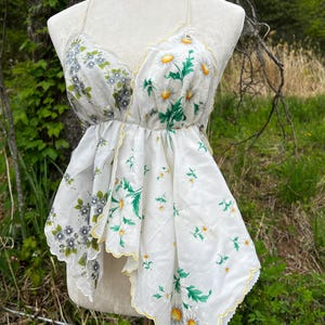 May include: A white halter top made from vintage handkerchiefs. The top features a floral print with daisies and other flowers in shades of yellow, green, and gray. The edges are scalloped and trimmed with yellow. The top is displayed on a mannequin.