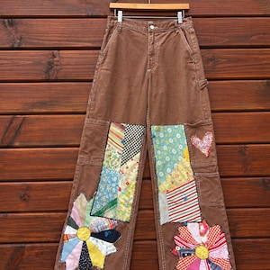 May include: Brown corduroy pants with colorful patchwork flower and heart appliqués. The wide-leg pants are hanging on a wooden hanger against a brown wooden background. The patchwork includes various patterns and colors.