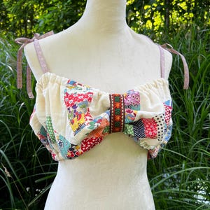 May include: A cropped top made from a patchwork of various colorful floral and geometric fabric squares. The garment features a gathered neckline, tie-up shoulder straps in a light mauve floral print, and a central vertical trim with orange flowers.