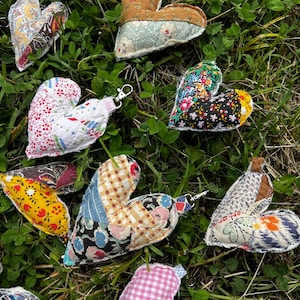 Puede incluir: Una colección de adornos en forma de corazón de tela hechos a mano en varios patrones y colores, incluyendo diseños florales, a cuadros y patchwork. Algunos corazones tienen llaveros adjuntos, sobre un fondo de hierba verde.