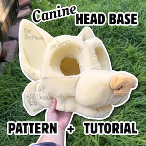 Pode incluir: Uma base de cabeça canina de espuma bege com orelhas grandes e pontiagudas, um focinho e órbitas oculares. A superfície tem etiquetas manuscritas, incluindo Ear Bottom, Up Cheek e Fluff Cheek Top. O texto Canine HEAD BASE PATTERN + TUTORIAL está sobreposto.