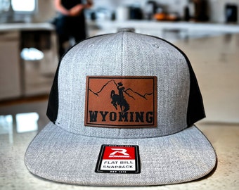 May include: Gray and black baseball cap with a brown leather patch that says "WYOMING" and a silhouette of a cowboy riding a horse. The patch is on the front of the cap. The cap has a black mesh back and a flat bill. The cap has a red and black label that says "FLAT BILL SNAPBACK" on the back.