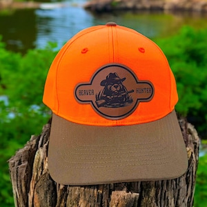 May include: An orange baseball cap with a brown brim. The cap features a leather patch with a beaver wearing a hat and holding a rifle, with the words "BEAVER HUNTER" in a banner.
