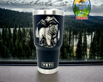 May include: A black Yeti tumbler with a laser engraved design of a bear walking through a mountain scene. The text "Wind River Outpost" is visible in the background.