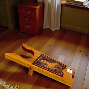 May include: A wooden boot jack with a decorative, dark brown inlay featuring a figure. The boot remover is made of light-colored wood and is designed to help remove boots. It is resting on a hardwood floor.