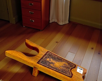 May include: A wooden boot jack with a decorative, dark brown inlay featuring a figure. The boot remover is made of light-colored wood and is designed to help remove boots. It is resting on a hardwood floor.