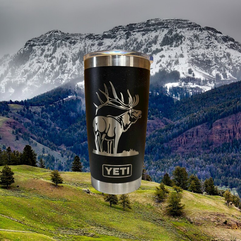 May include: A black Yeti tumbler with a silver lid and a laser-etched design of an elk on a mountain background. The tumbler has the word "YETI" printed on the bottom.