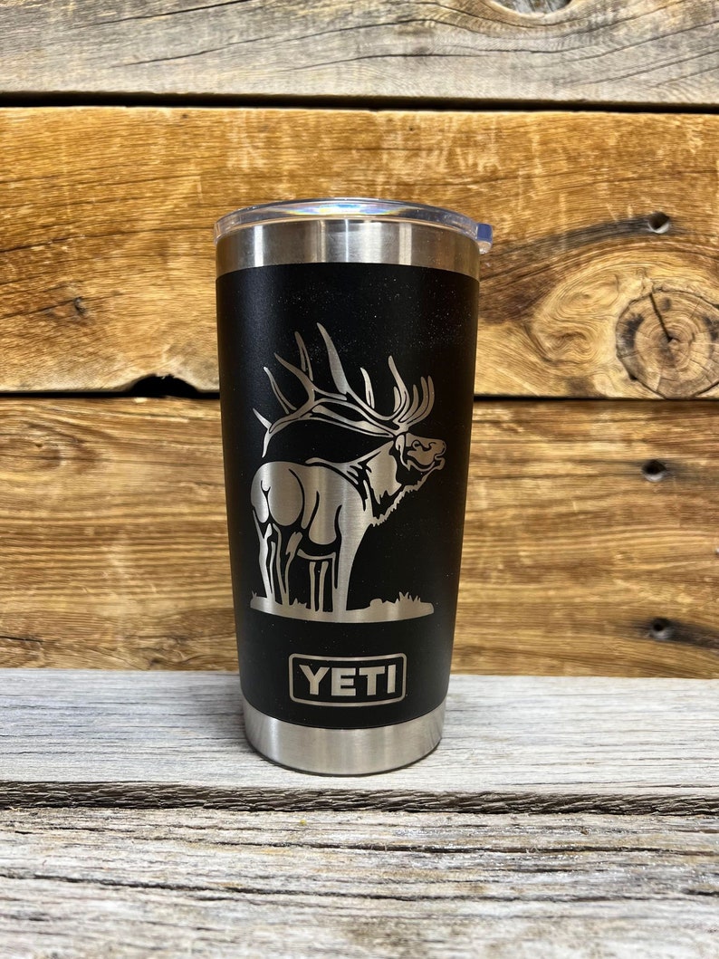 May include: A black Yeti tumbler with a laser-etched design of an elk. The tumbler has a stainless steel exterior and a silver lid. The word "YETI" is printed on the front of the tumbler.