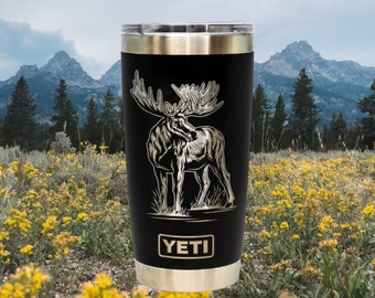 May include: Black Yeti tumbler with a silver lid and a laser-etched design of a moose on a black background. The text "YETI" is printed in silver on the bottom of the tumbler.