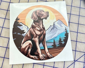 May include: A brown and white dog sits in a mountain landscape with a sunset in the background. The dog is looking to the right of the image. The mountains are in the background and have snow on them. There are trees in the foreground.