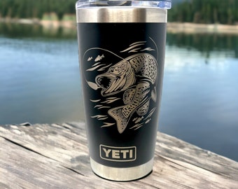 May include: A black YETI tumbler with a gold-colored graphic of a fish jumping out of the water. The tumbler has a stainless steel lid and base.