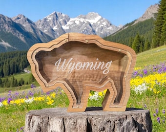 May include: A wooden bison-shaped decorative box with the word "Wyoming" in white script. The box is made of light brown wood and sits on a tree stump. The background features mountains, trees, and wildflowers.