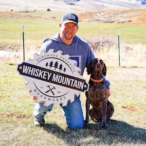 May include: A man in a grey hoodie and a black baseball cap is kneeling in a grassy field with a dog. He is holding a white circular sign with black text that reads "Handmade Products - Whiskey Mountain - Est. 2009 - Woodworks".