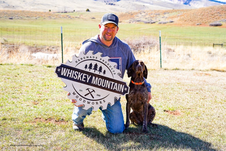 May include: A man in a grey hoodie and a black baseball cap is kneeling in a grassy field with a dog. He is holding a white circular sign with black text that reads "Handmade Products - Whiskey Mountain - Est. 2009 - Woodworks".