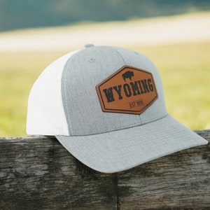 May include: A gray and white trucker hat with a brown leather patch that says "WYOMING EST 1890" and features a silhouette of a buffalo.