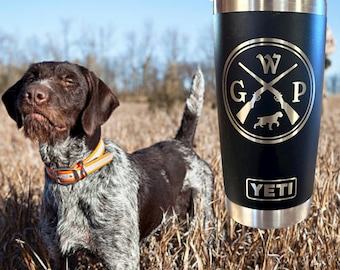 May include: A black Yeti tumbler with a silver lid and a laser engraved design of crossed shotguns with the letters "WGP" and a small dog silhouette. The tumbler is sitting on a bed of dry grass with a brown and white dog in the background.