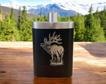May include: Black stainless steel flask with an engraved elk design and the word "YETI" on the bottom. The flask is sitting on a wooden surface.