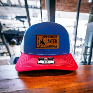 May include: A blue, white, and red trucker hat with a brown patch that says "Wyoming Lander" and a silhouette of a cowboy riding a horse. The hat is made by Richardson 112.