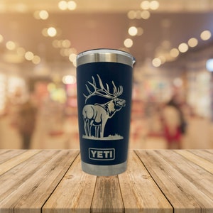 May include: A blue Yeti tumbler with a silver lid and a laser-etched design of an elk. The tumbler has the word "YETI" printed on the bottom.