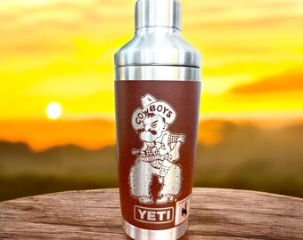 May include: A stainless steel cocktail shaker with a brown leather wrap featuring a vintage cartoon cowboy graphic and the text "COWBOYS" and "YETI".