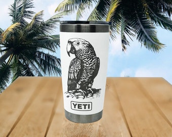 May include: A black and white illustration of a parrot on a white tumbler with the word "YETI" printed on the bottom. The tumbler is sitting on a wooden surface.