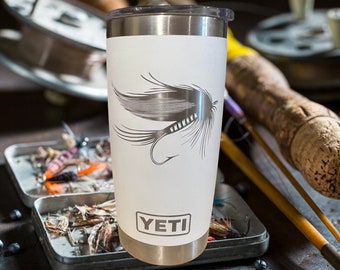 May include: White Yeti tumbler with a silver lid and a black and white graphic of a fly fishing lure. The tumbler has the word "YETI" printed on the bottom.