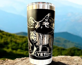 May include: A black Yeti tumbler with a silver lid and a laser-etched design of a wolf and mountains. The text "YETI" is printed on the front of the tumbler.