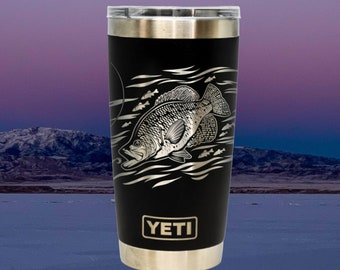 May include: Black Yeti tumbler with a silver lid and base. The tumbler features a laser-etched design of a crappie fish swimming in water. The word "YETI" is printed in gold on the front of the tumbler.