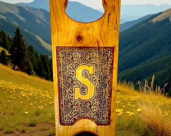 May include: A wooden boot jack with a decorative panel featuring the letter "S" in gold. The panel is framed by a brown border and intricate gold scrollwork. The boot jack is made of light-colored wood.
