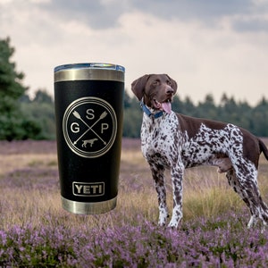 May include: A black Yeti tumbler with a gold logo that says "SGP" with a dog silhouette and crossed arrows. The tumbler is sitting on a bed of purple flowers with a German Shorthaired Pointer dog standing in the background.