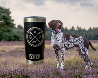 May include: A black Yeti tumbler with a gold logo that says "SGP" with a dog silhouette and crossed arrows. The tumbler is sitting on a bed of purple flowers with a German Shorthaired Pointer dog standing in the background.