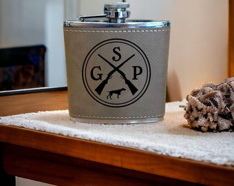 May include: A gray leather flask with a silver top and a black design of crossed shotguns, a dog, and the letters "GSP".