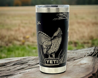 May include: A black Yeti tumbler with a laser-etched design of a ruffed grouse. The tumbler has a silver lid and base and the word "YETI" is printed on the side.