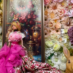 Könnte beinhalten: Eine Porzellanpuppe in einem rosa und floralen Kleid mit einer Krone und einer Perlenkette. Die Puppe steht vor einem gerahmten Gemälde mit Blumen und einer Vase. Der Hintergrund ist eine Wand aus künstlichen Blumen.