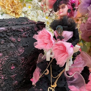 Könnte beinhalten: Eine schwarz-rosa Puppe mit einem rosa Hut und einem schwarz-weißen Kleid. Die Puppe trägt eine goldene Halskette mit Halbmond- und Herzanhängern. Die Puppe steht vor einer schwarz-rosa verzierten Schachtel.