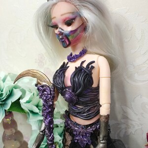 May include: A doll with white hair and a black crown wears a black and purple outfit with a crescent moon-shaped staff. The doll has a painted face with rainbow colors.