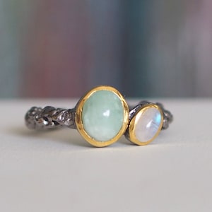 Può includere: Un anello d'argento con una fascia intrecciata e due pietre preziose. La pietra più grande è un'acquamarina verde chiaro, e la pietra più piccola è una pietra di luna bianca.