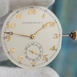 May include: A vintage watch face with gold numerals and hands, held by a gloved hand. The watch face is cream-coloured with a small seconds dial and the text "TATANNER WATCH CO". The watch has a gold crown.