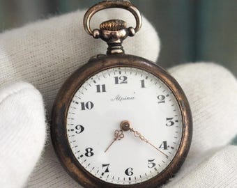 Puede incluir: Un pequeño reloj de bolsillo antiguo con esfera blanca y números negros. El reloj tiene una caja de color bronce y un pequeño lazo en la parte superior. Las manecillas son doradas. El reloj se sostiene en una mano enguantada de blanco.