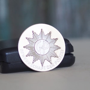 May include: A silver coin with a starburst design and the text "GEWERKSCHAFT DER POLIZEI" around the edge. The center of the coin features the letters "GP".