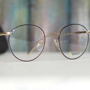 May include: Round eyeglasses with gold-coloured frames and clear lenses. The brand name "Vasco" is printed on the right lens.